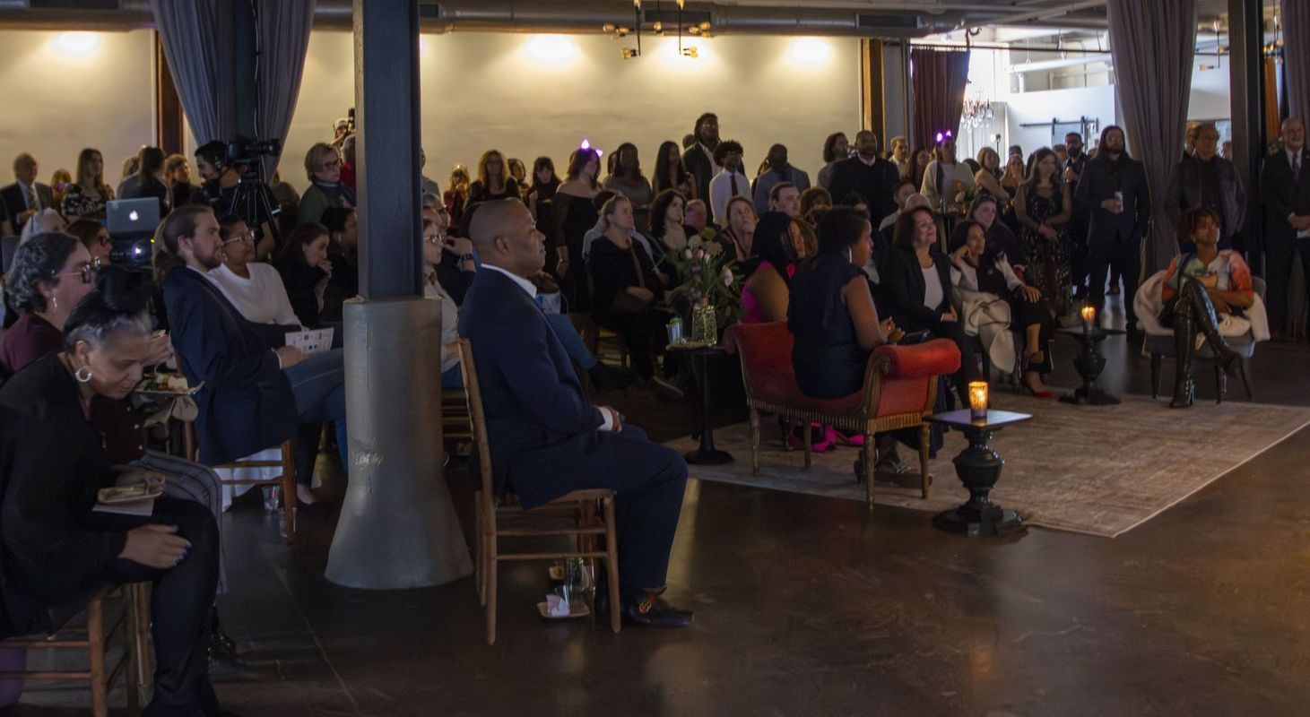 Audience seated, listening to speaker. Lit, large event space. Dark suits, casual attire.
