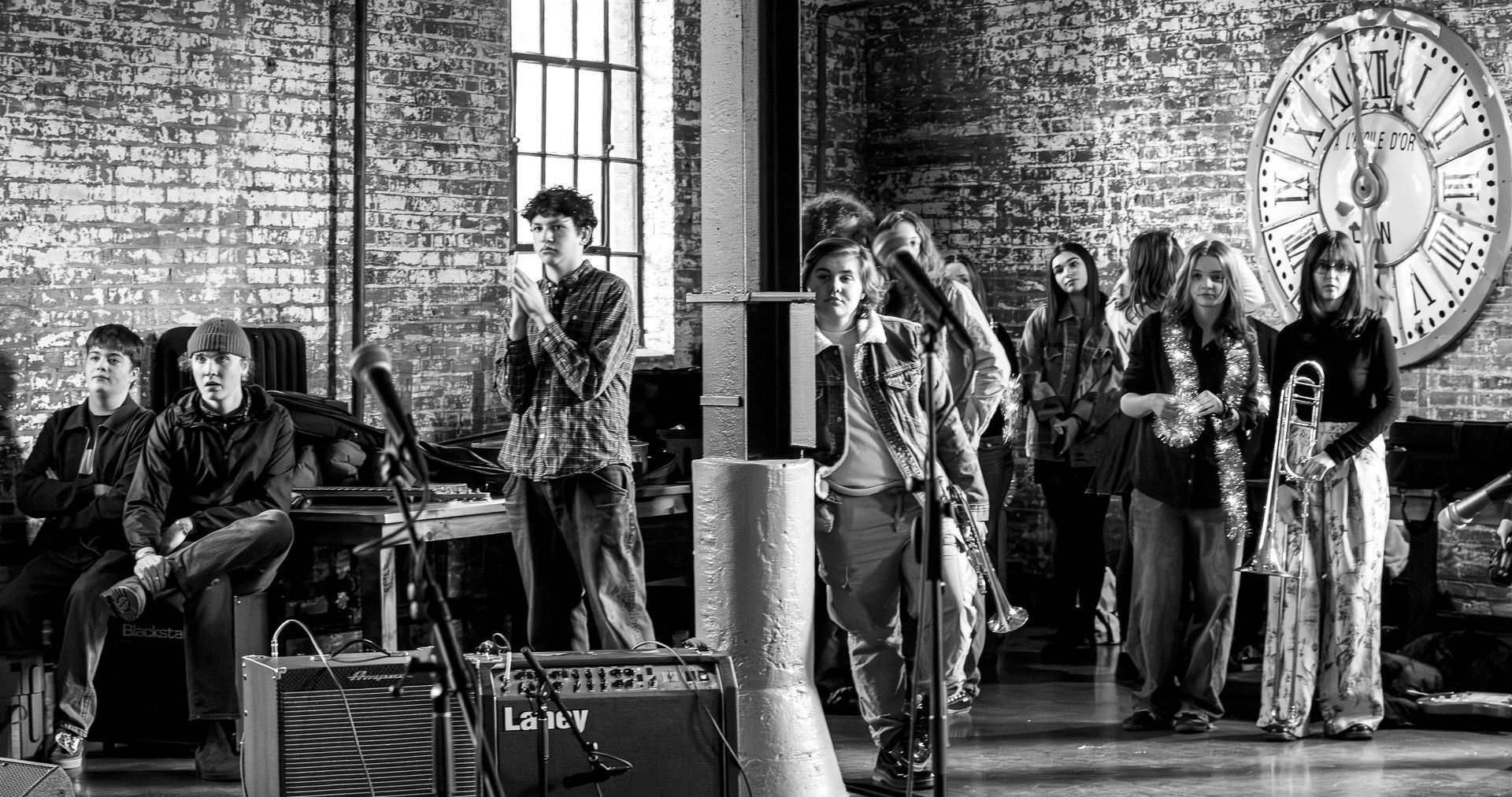 Band performing in a brick-walled room with a large clock. Musicians playing instruments and singing, in grayscale.