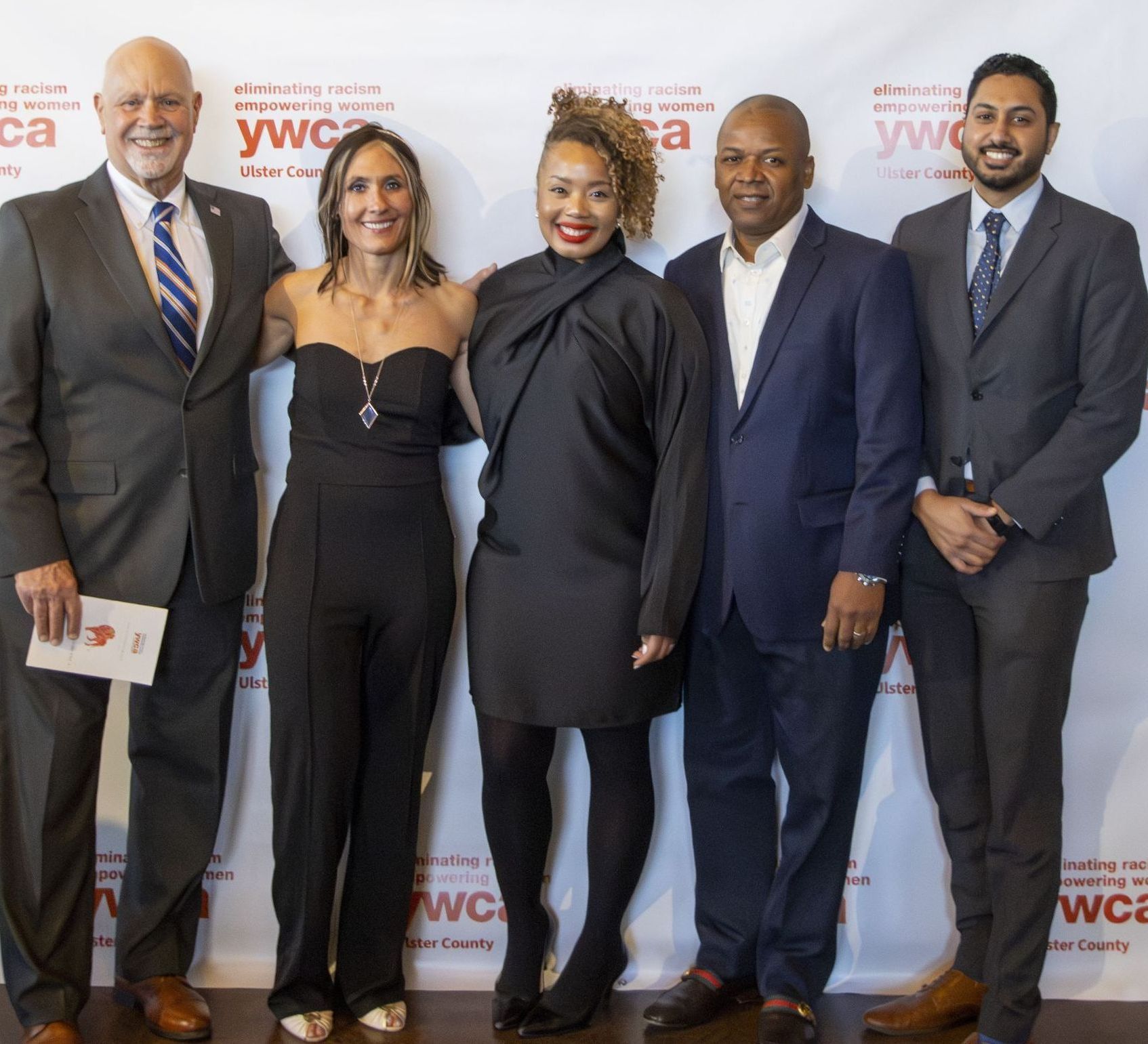 Five people posing at an event; a man in a suit and tie, a woman in black jumpsuit, and three other people smiling.