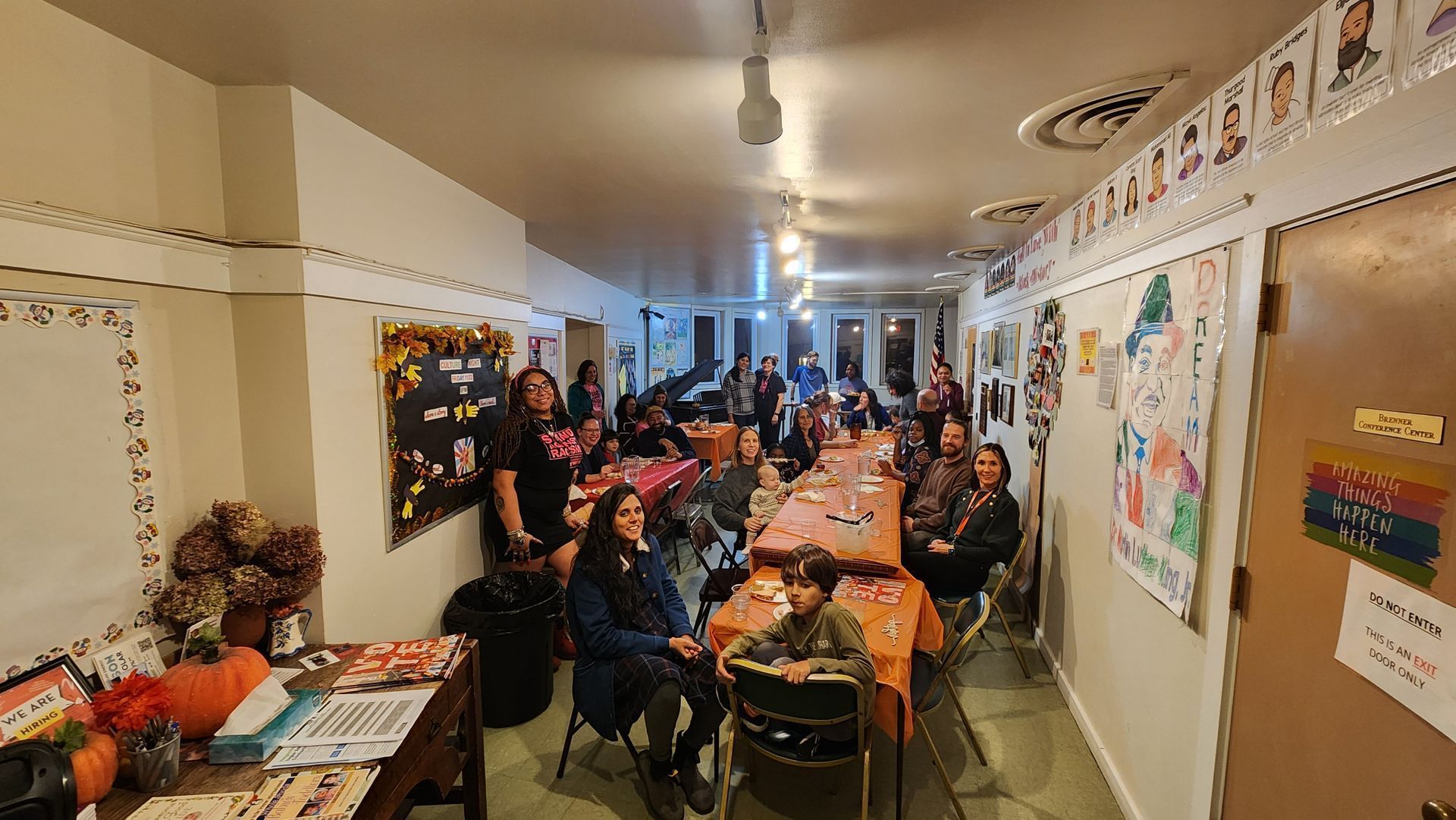 A long room with a group of people at a table, some smiling. Decorations, windows, and lights are visible.