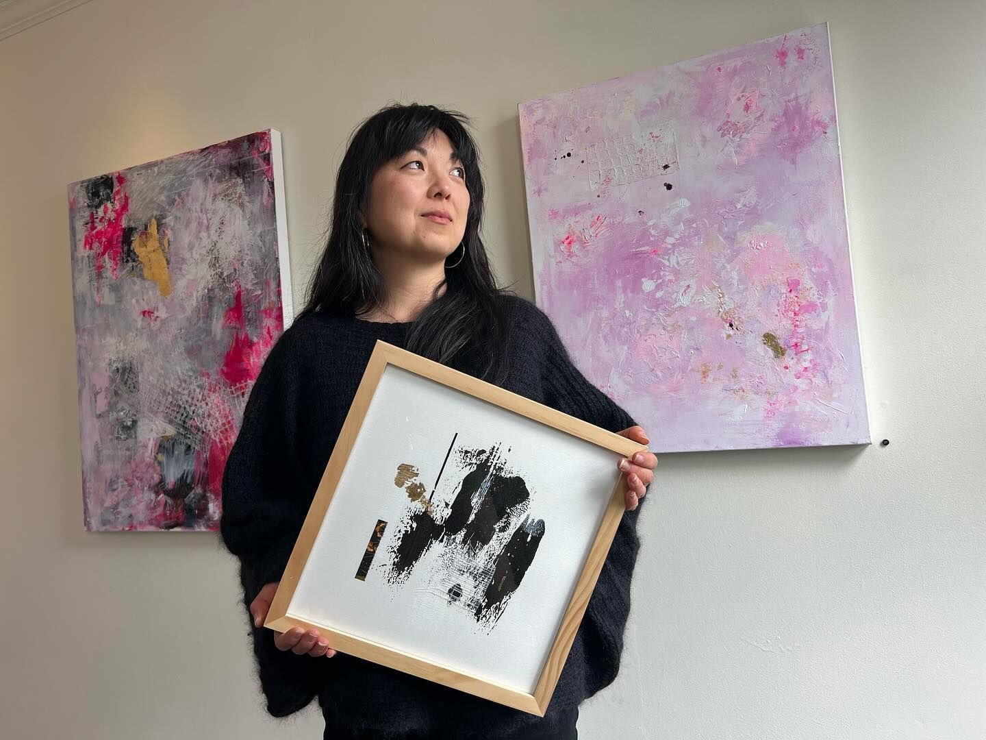 Woman holding art, standing in front of her abstract paintings. Paintings have pink, gray, and gold colors.