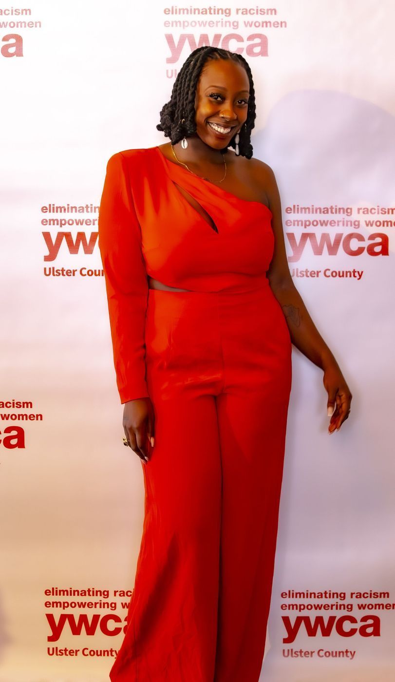 Woman in red one-shoulder jumpsuit smiles at YWCA event, posing against a red and white backdrop.
