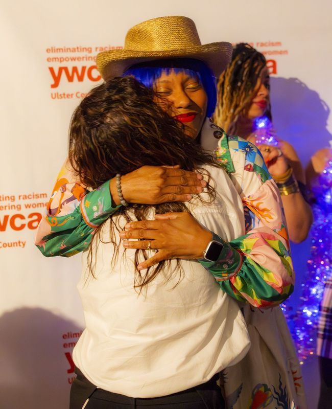 Two women embrace, one in floral shirt, straw hat, blue wig; YWCA event.