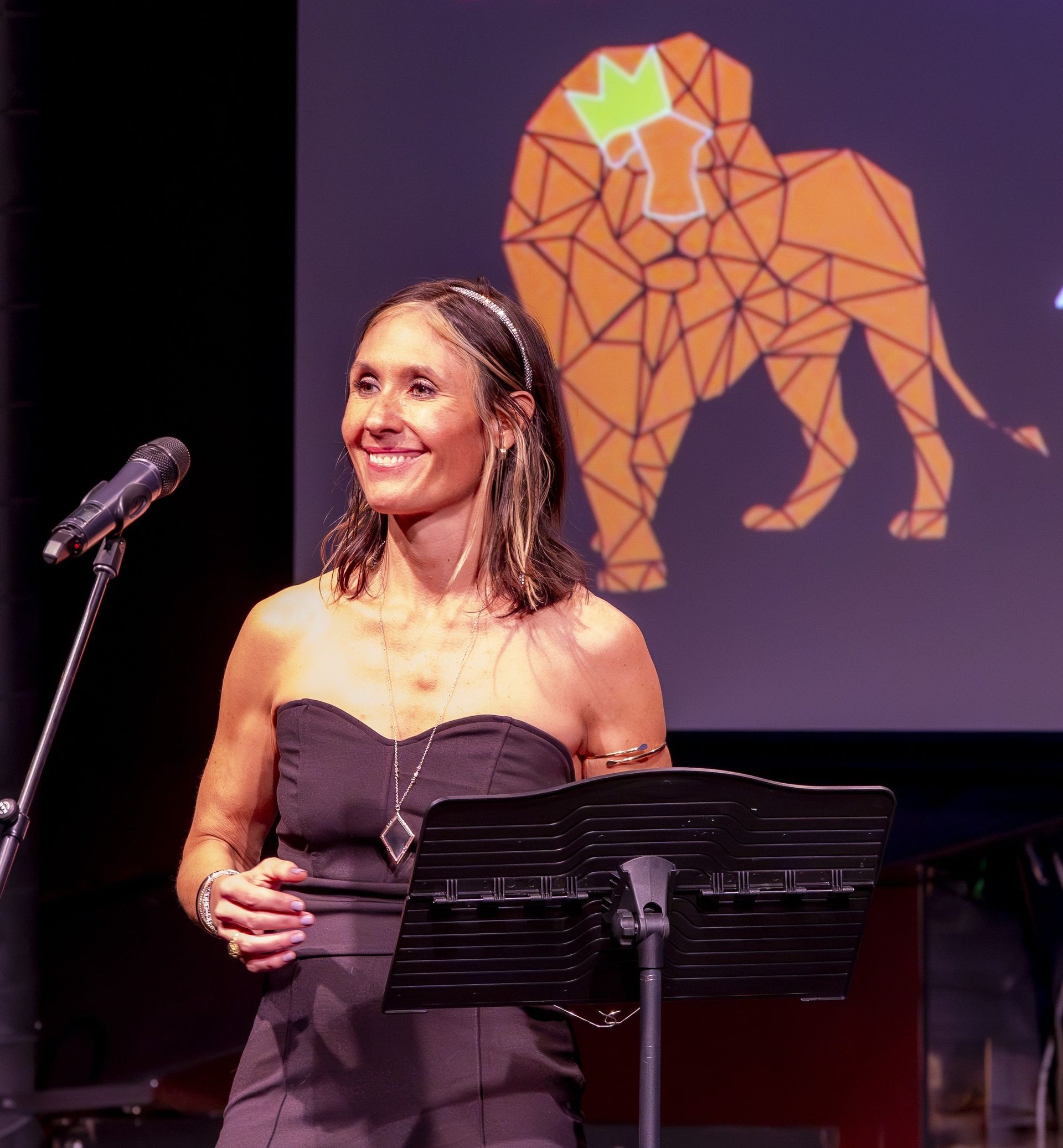 Woman in a black strapless dress speaks at a podium on stage; a lion logo in the background.