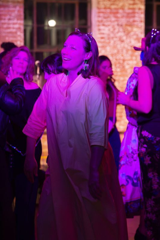 Woman dances at a party, lit by pink light. She wears a yellow dress and a tiara, smiling. Other people are blurred in the background.