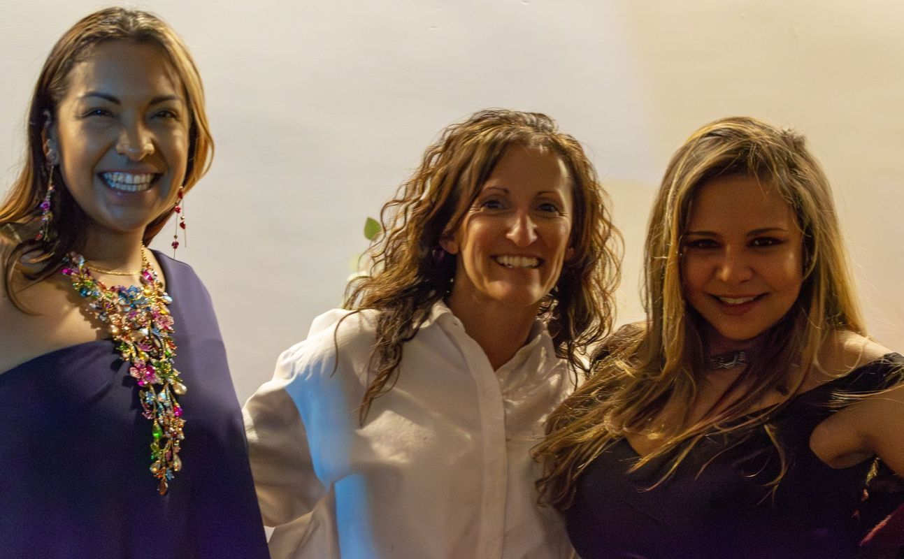 Three smiling women posing indoors. One wears a colorful necklace, another a white shirt, and the third a dark dress.