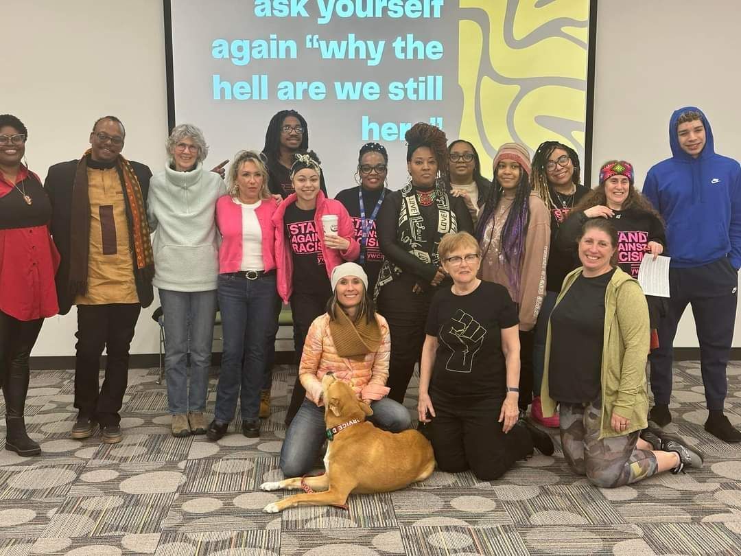 Group of diverse people pose, some hugging, in front of a screen with text and a design. A deer sits in the center.