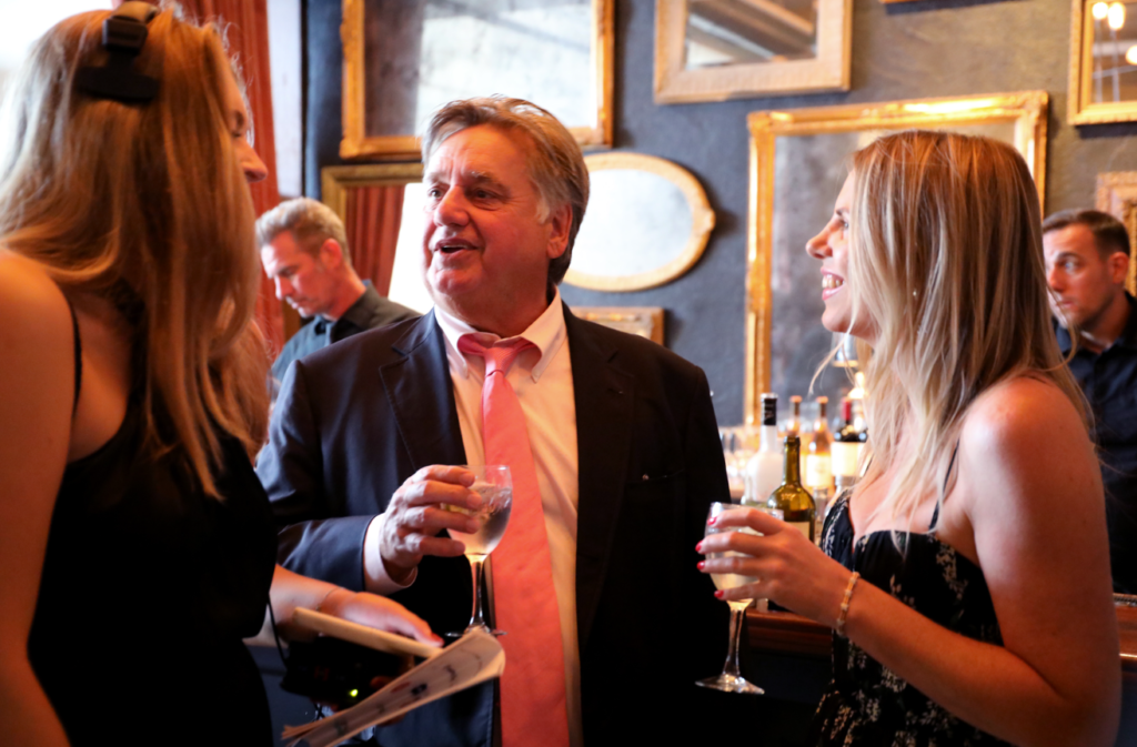 Man in suit talking to two women, holding glasses in a room with mirrors; others in background.