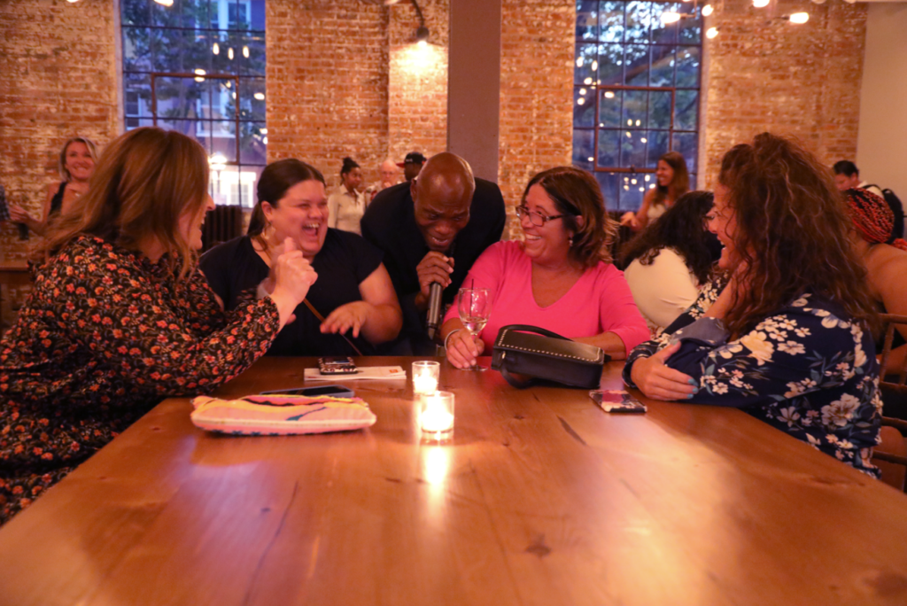Group of women at a table with a man singing; warmly lit restaurant.