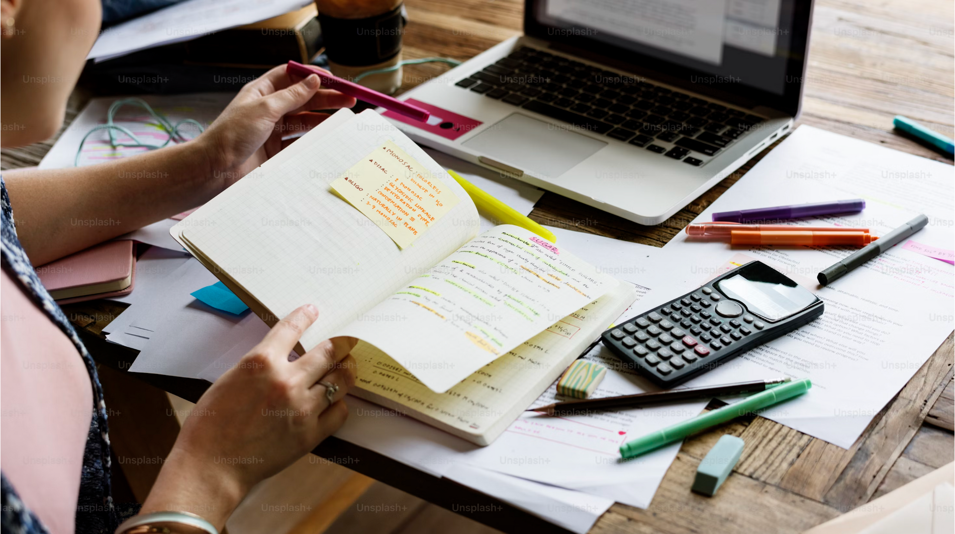 Person looking at a notebook on a cluttered desk with laptop, calculator, pens, and papers.