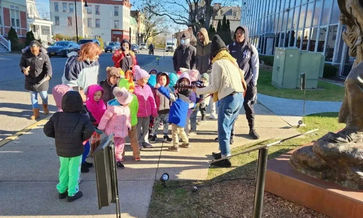 Children in colorful coats on a field trip, looking at a sculpture in front of a building.