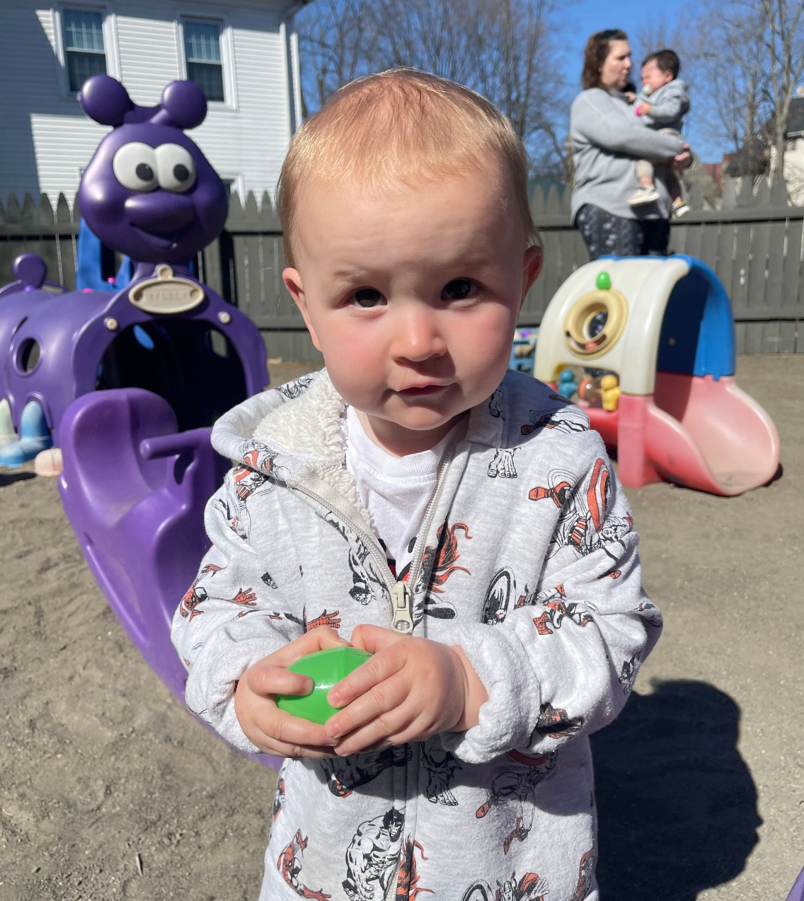 Blonde baby in grey onesie holding a green toy, standing in a playground, purple slide and a woman holding a baby in the background.