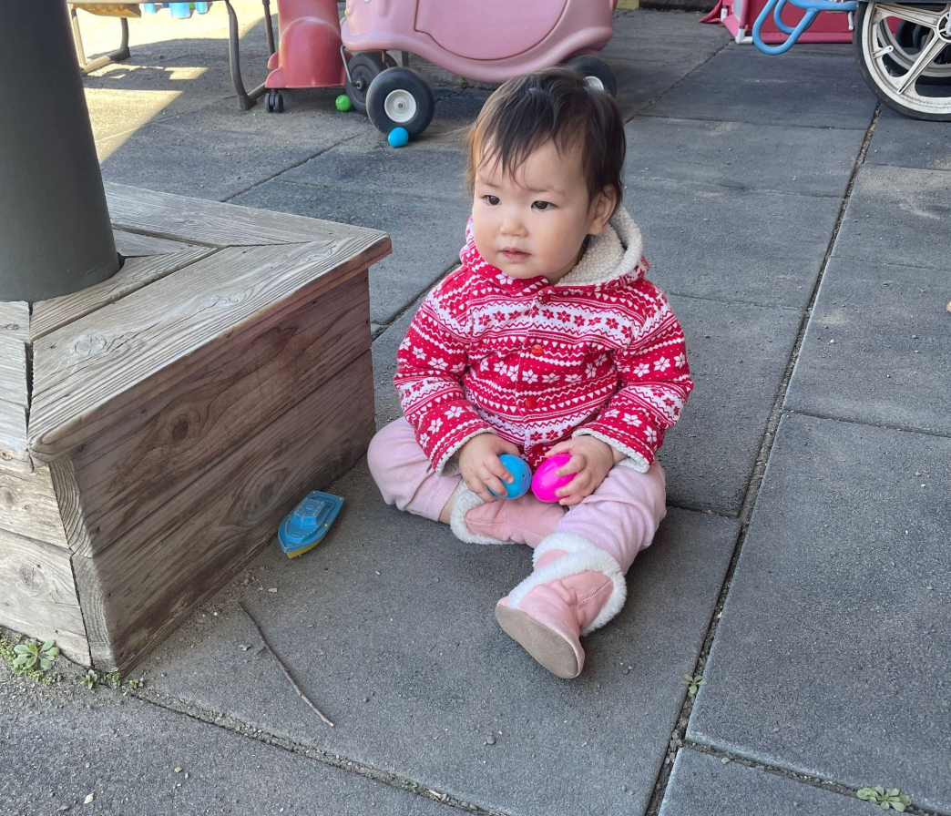 Baby sitting on ground wearing a red sweater, holding toys, with playground in the background.