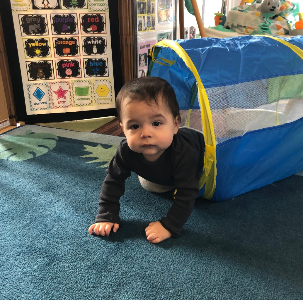 Baby crawls out of a blue and yellow play tunnel on a blue carpet; looking at the camera.