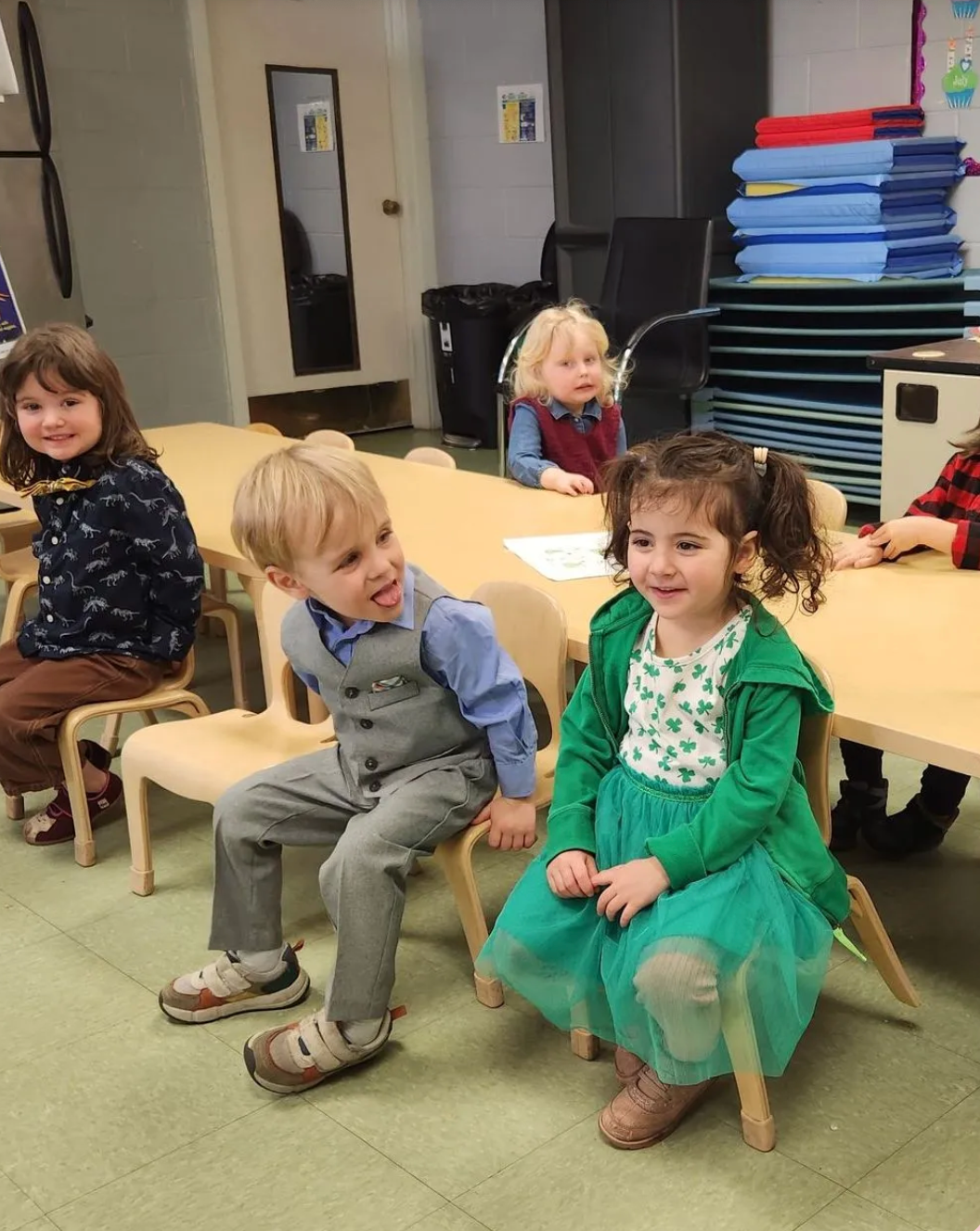 Children sitting at a table in a classroom. One boy sticks his tongue out, another smiles.