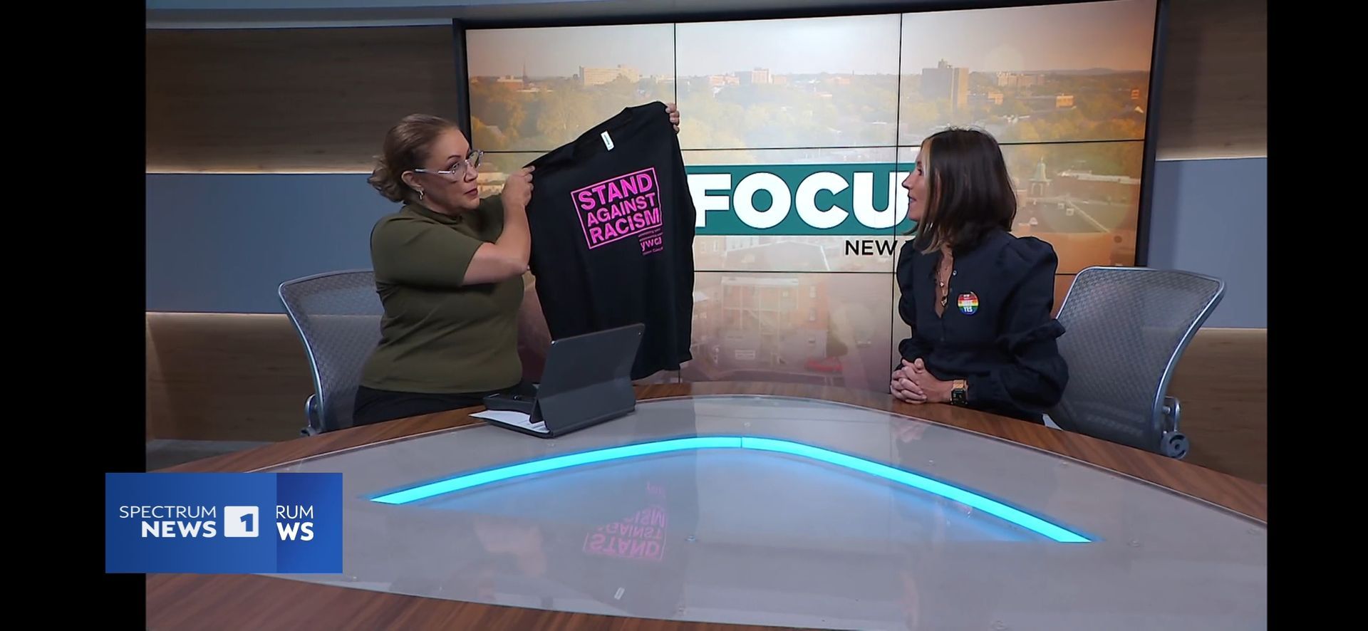 Two women in a news studio, one holding a black sweatshirt with pink text, the other looking on.