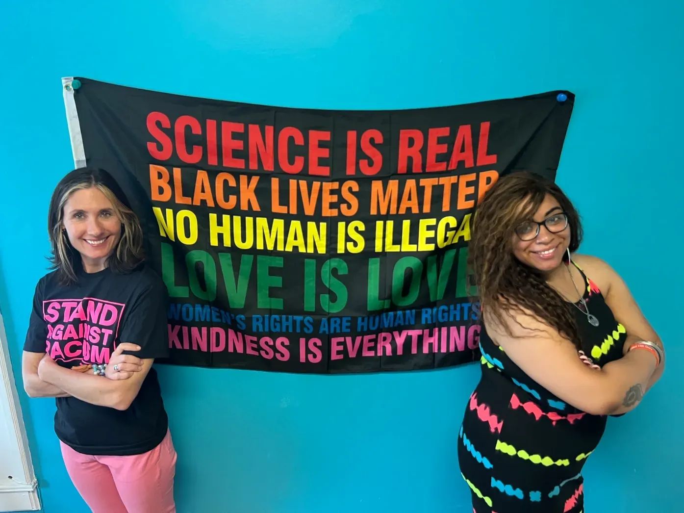 Two women stand with arms crossed in front of a banner with inclusivity messages. The banner is rainbow-colored on a black background.