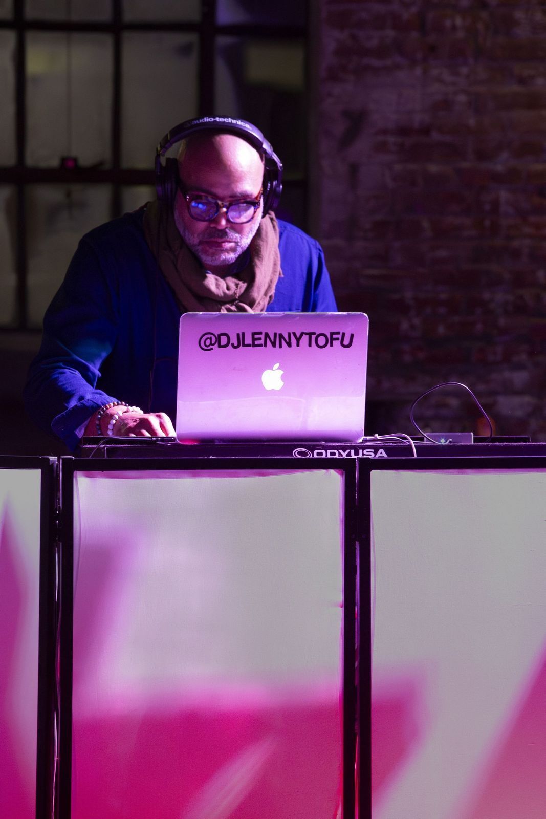 DJ wearing headphones, laptop on a stand, performing at an event with pink and white lighting.