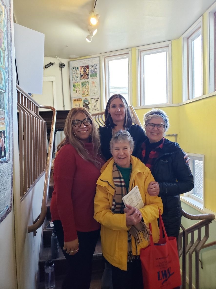 Four women smiling together on a staircase; one wears a yellow coat, one a red sweater, and two have jackets.