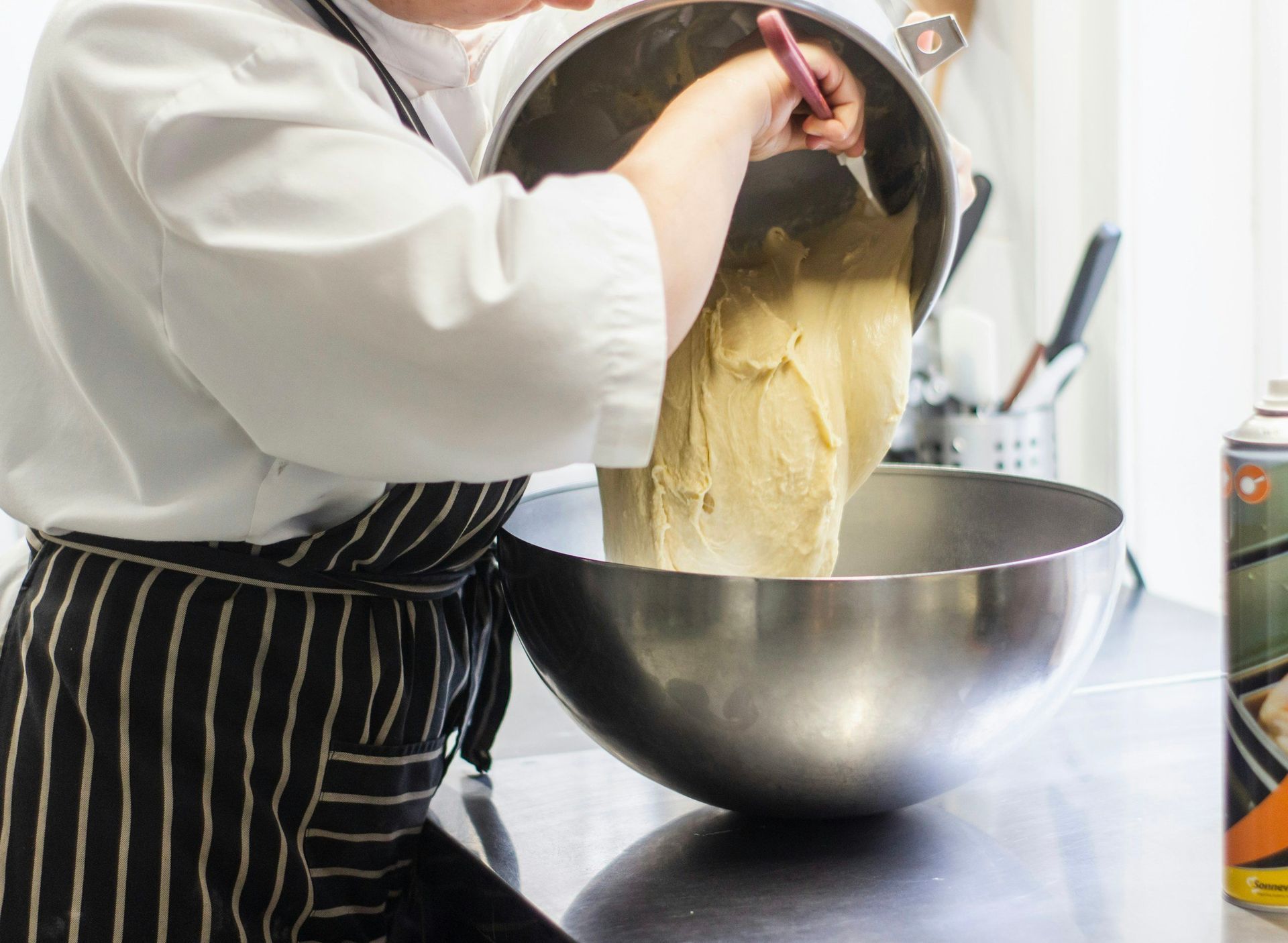 Chef in white uniform and striped apron pouring dough from a metal bowl into another metal bowl on a kitchen counter.