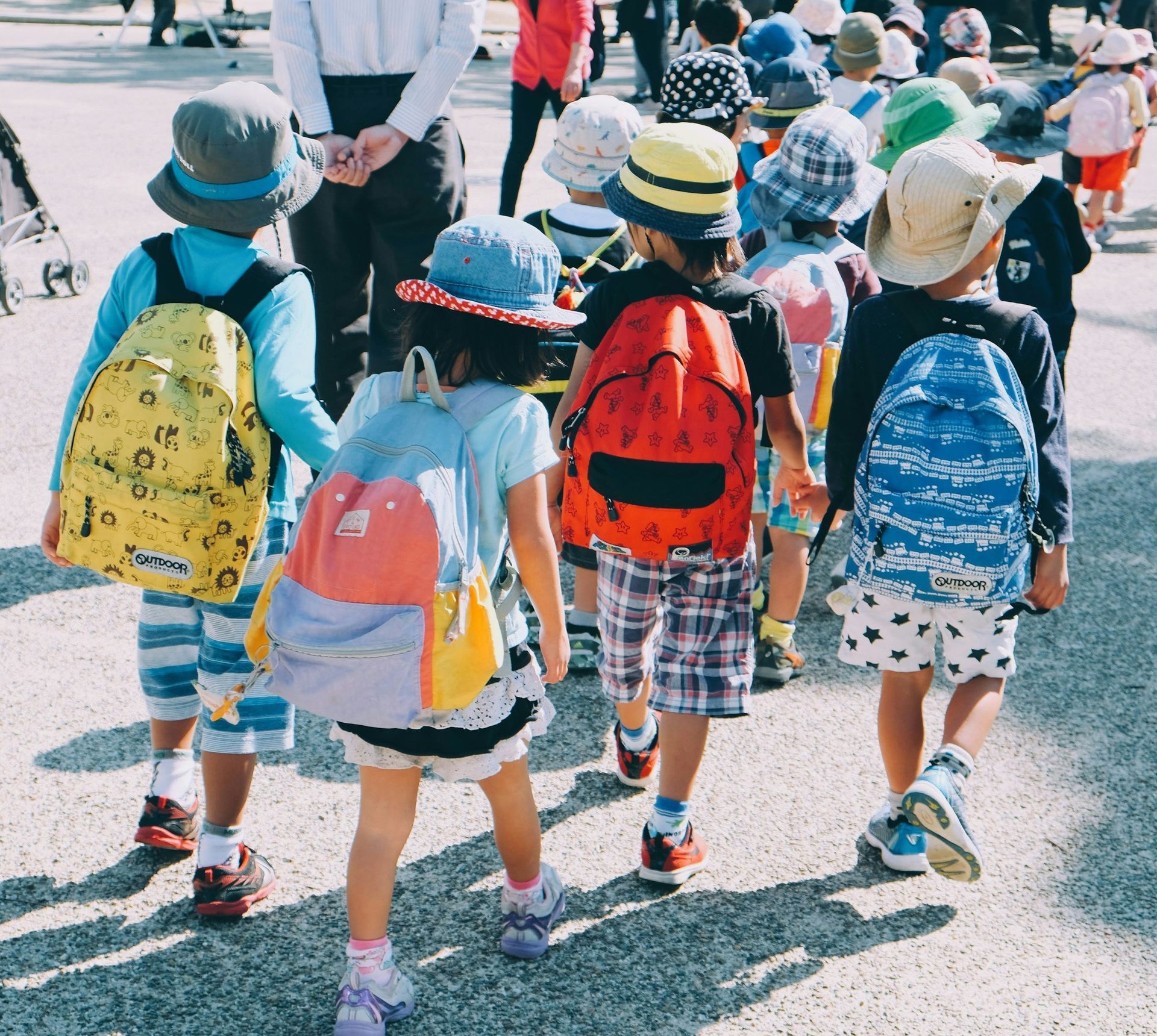 Children with backpacks walk in a group, some holding hands. Sunny day.