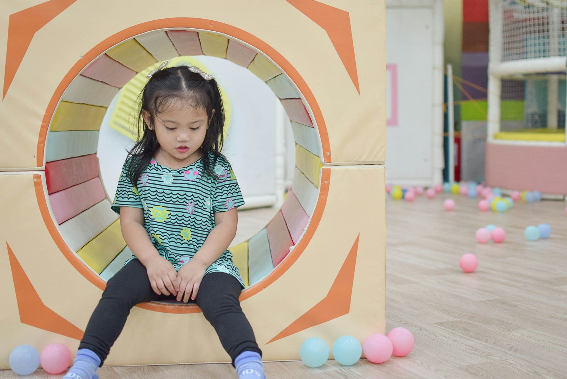 Young girl sits inside a soft play tunnel, looking down. Colorful balls scattered around.