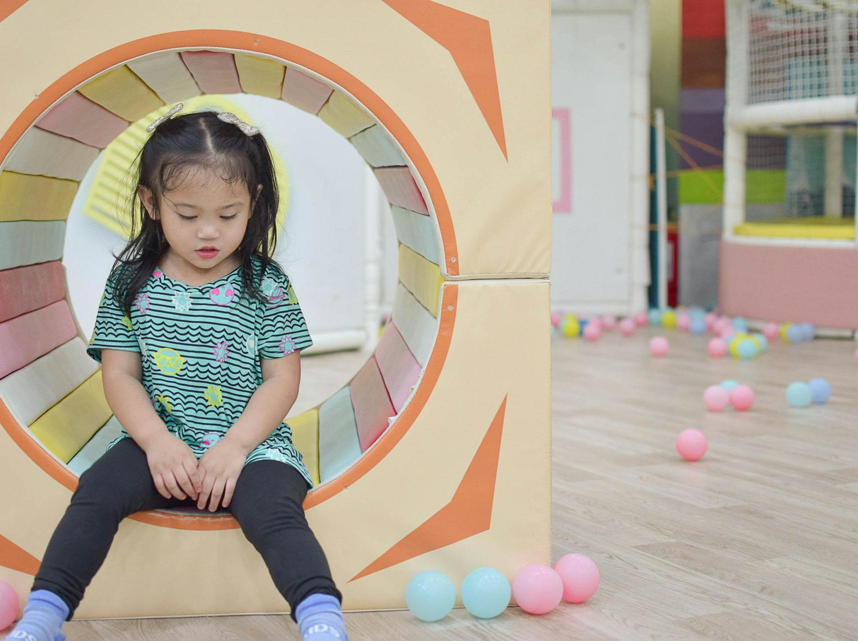Young girl sits inside a soft play tunnel, looking down. Colorful balls scattered around.