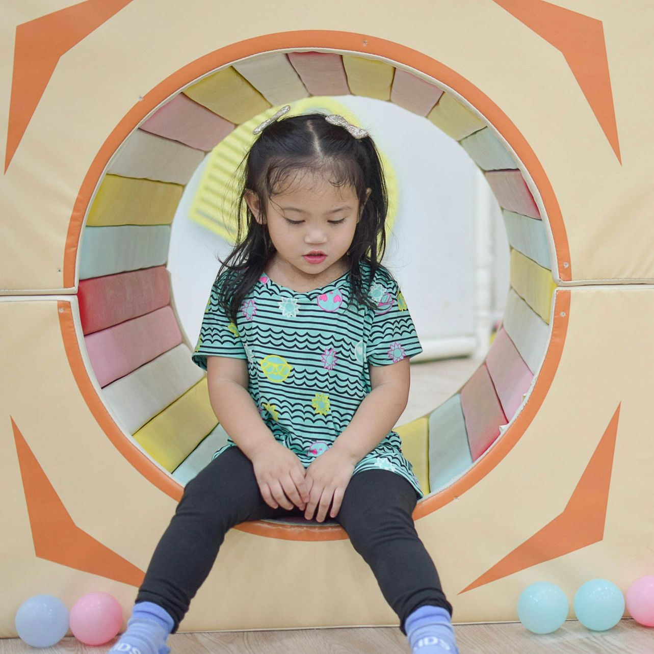 Young girl sits inside a soft play tunnel, looking down. Colorful balls scattered around.