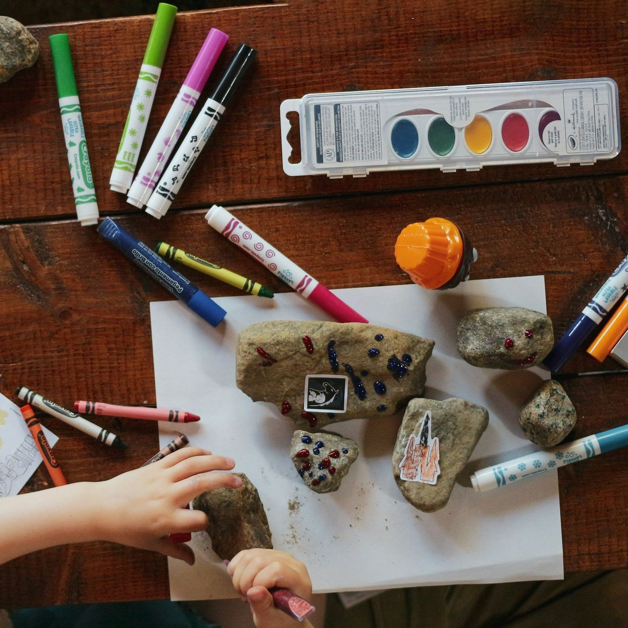 Child drawing on rocks with markers and crayons on a wooden table.