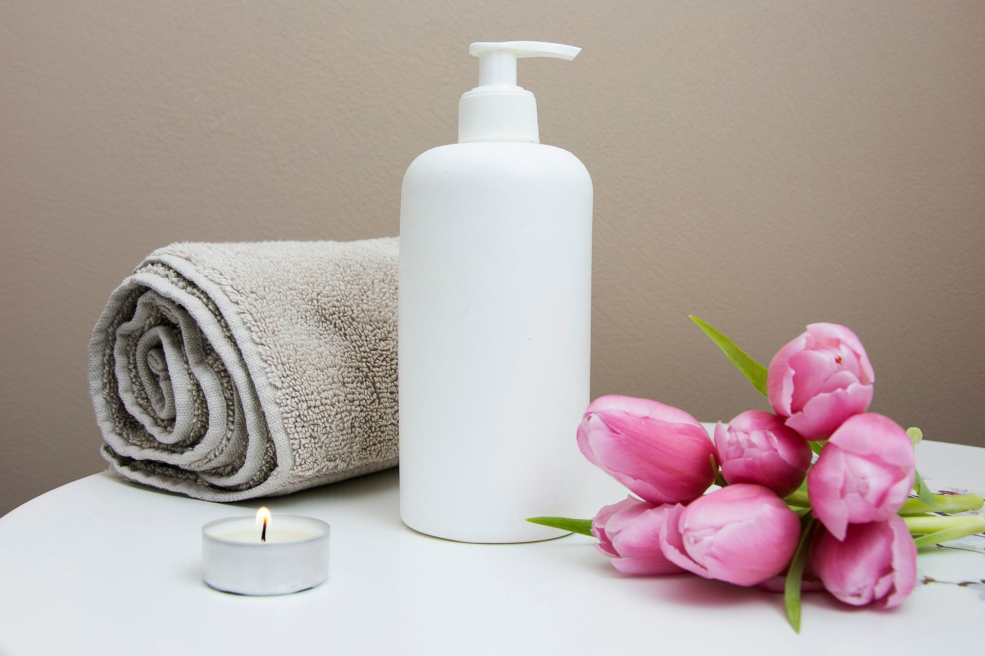 White lotion bottle, rolled towel, pink tulips, and lit candle on a white surface.