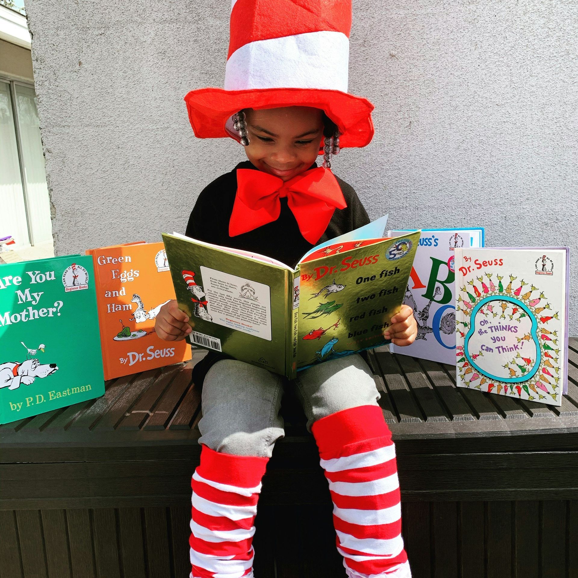 Child dressed as Cat in the Hat reading Dr. Seuss books, wearing red and white striped hat, bow tie, and leg warmers.