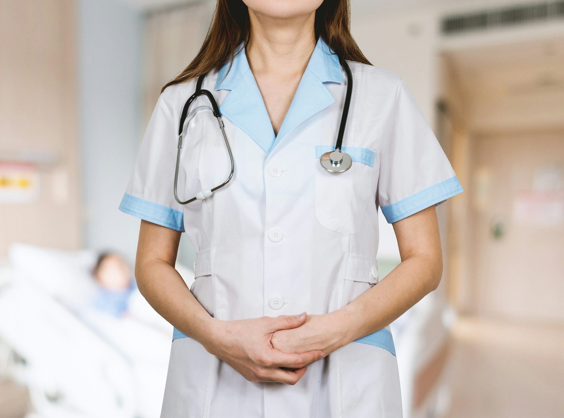 Nurse wearing a blue-trimmed white coat and stethoscope, standing in a hospital room.