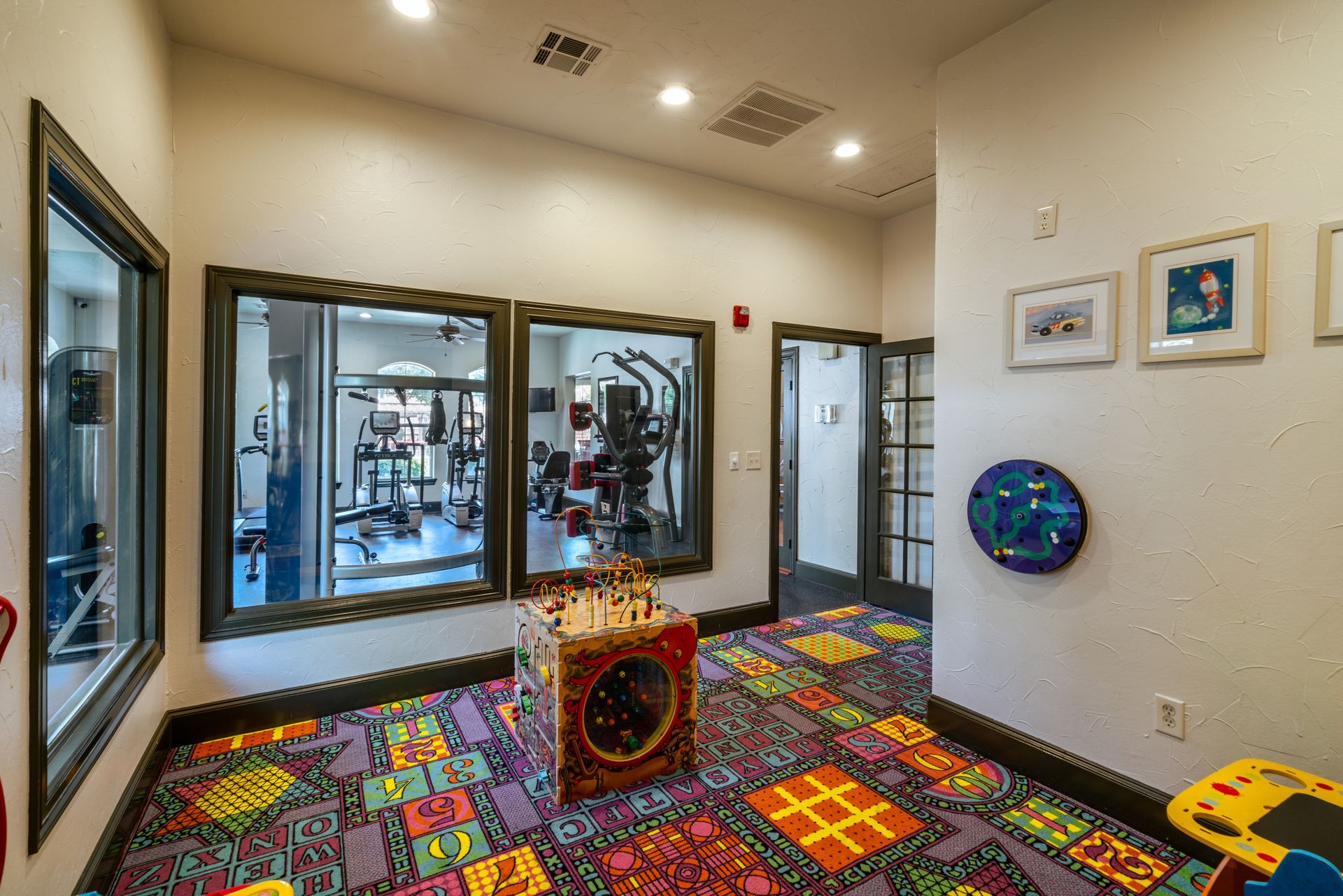 Hallway with colorful patterned carpet, windows looking into a gym, and a play area with toys.