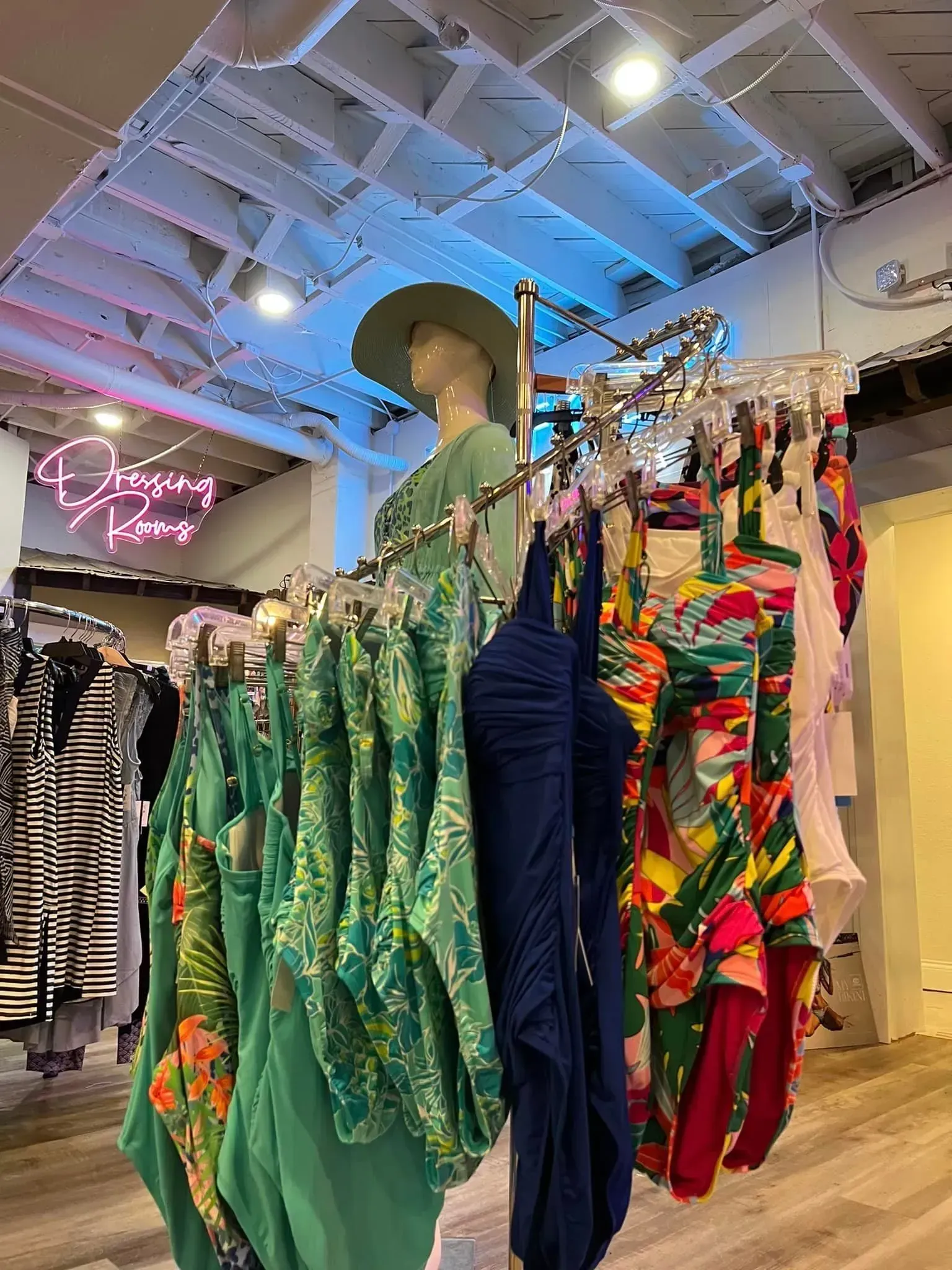 Clothing store interior with racks of colorful dresses and swimwear. A mannequin wearing a hat stands near a rack.