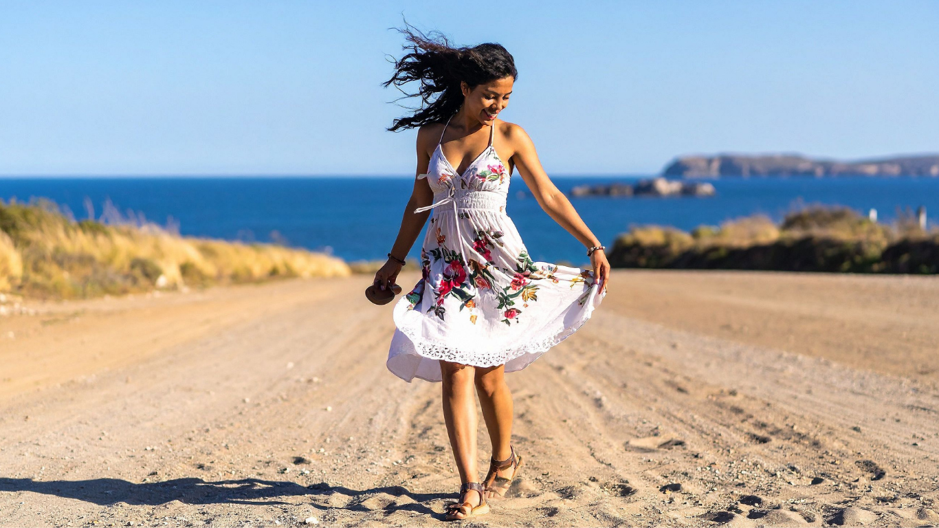 Woman in a white floral dress twirling on a dirt road, with the ocean in the background.