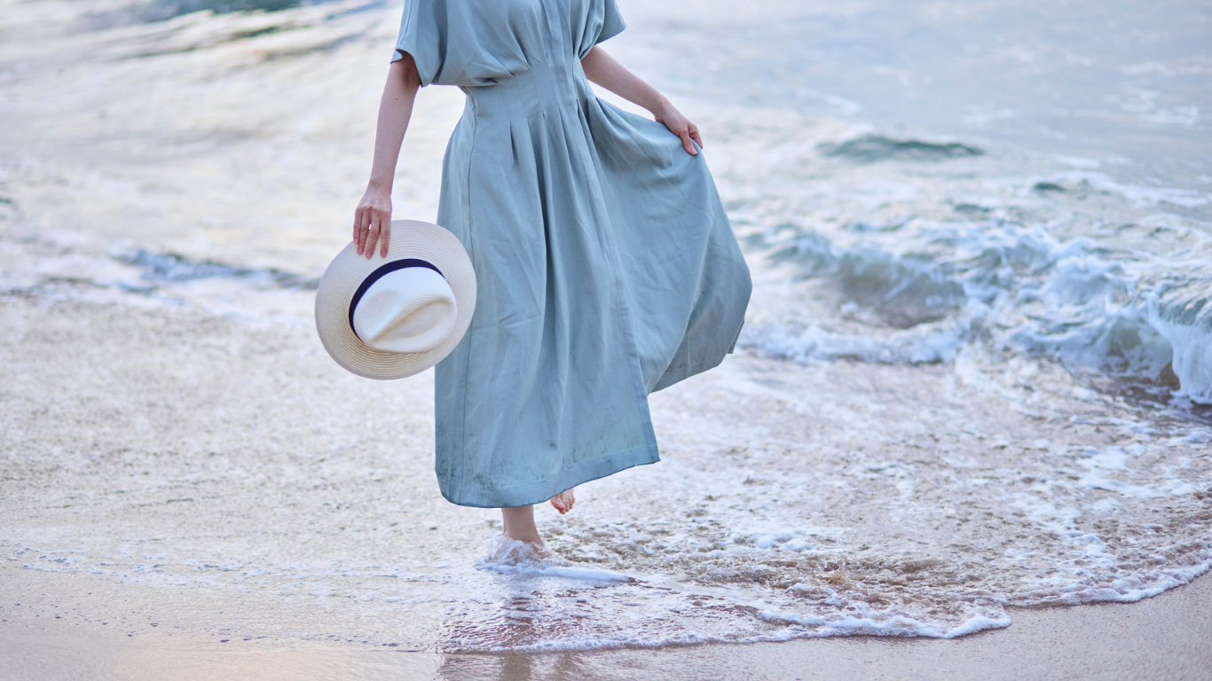 Woman in a blue dress stands in the ocean, holding a hat.