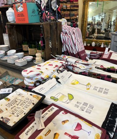 Kitchenware and gifts displayed on a shop table, including oven mitts, towels, and dishware with food and drink motifs.