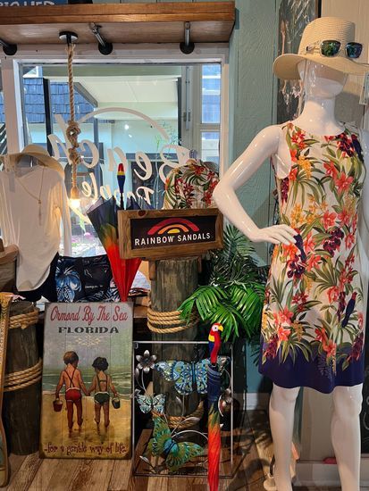 Mannequin in floral dress, beach-themed shop display. Includes sign, artwork, tropical decor, and window view.