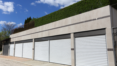 Row of white garage doors beneath a tall concrete wall with green hedging under a blue sky