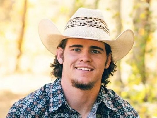 A young man wearing a cowboy hat and a floral shirt is smiling.