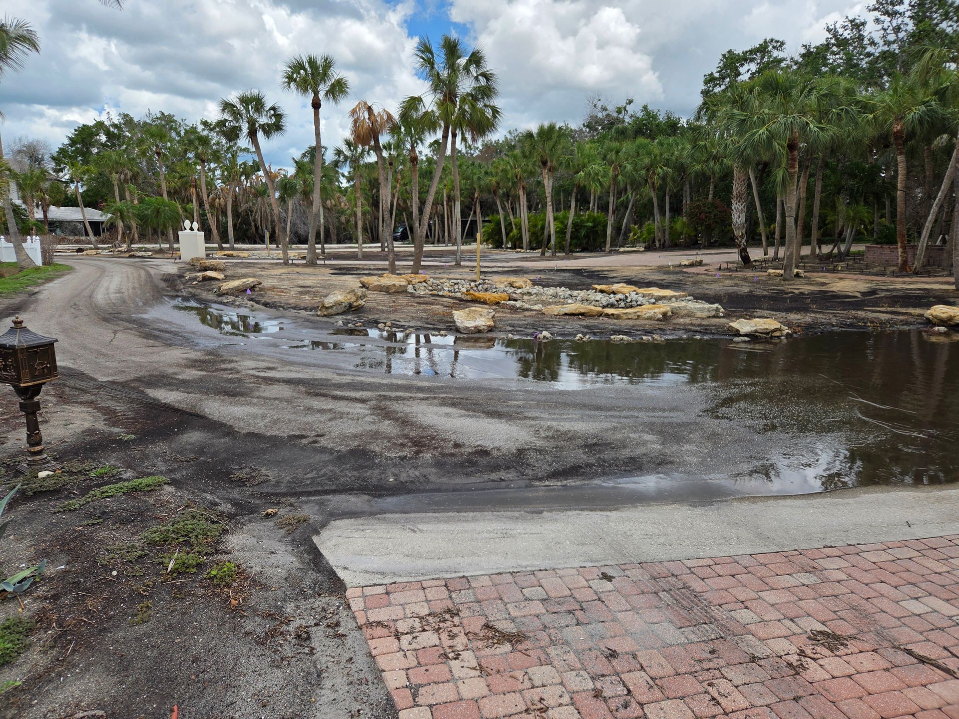 Puddle of water on dirt and brick surface with some trees in the background. Cloudy day.