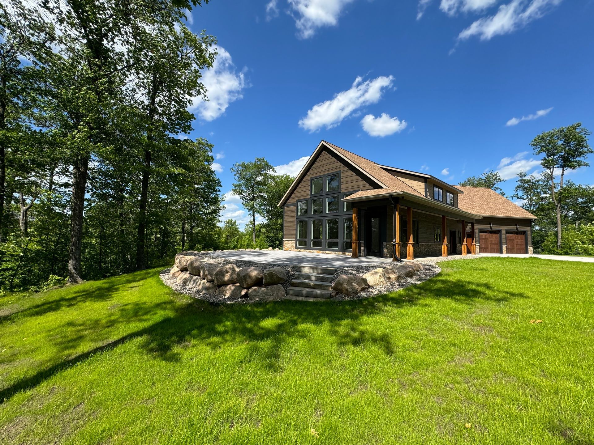 House with stone patio and garage on a grassy hill, surrounded by trees under a blue sky with clouds.