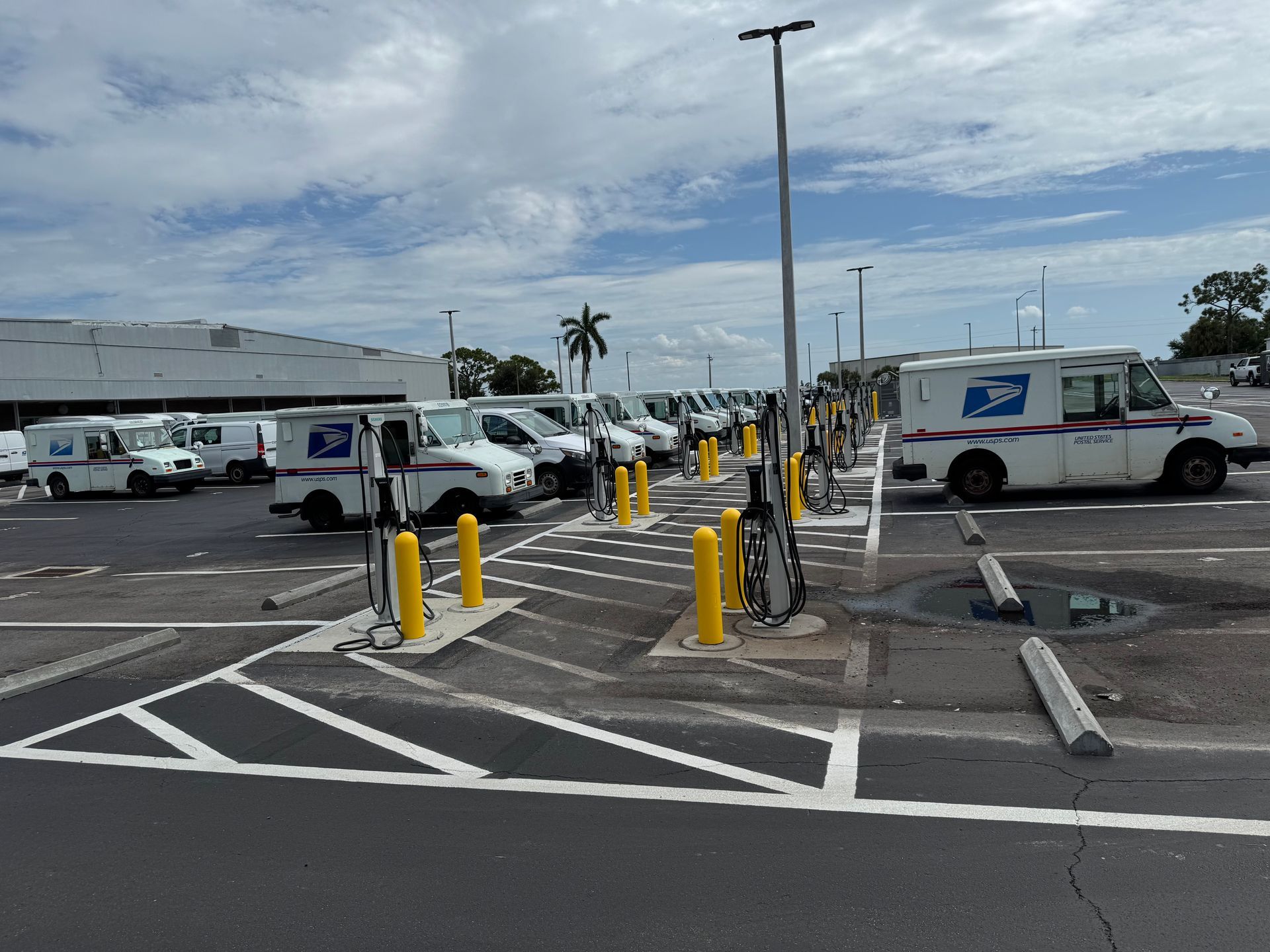 Postal service trucks parked at charging stations outside a building under a cloudy sky.