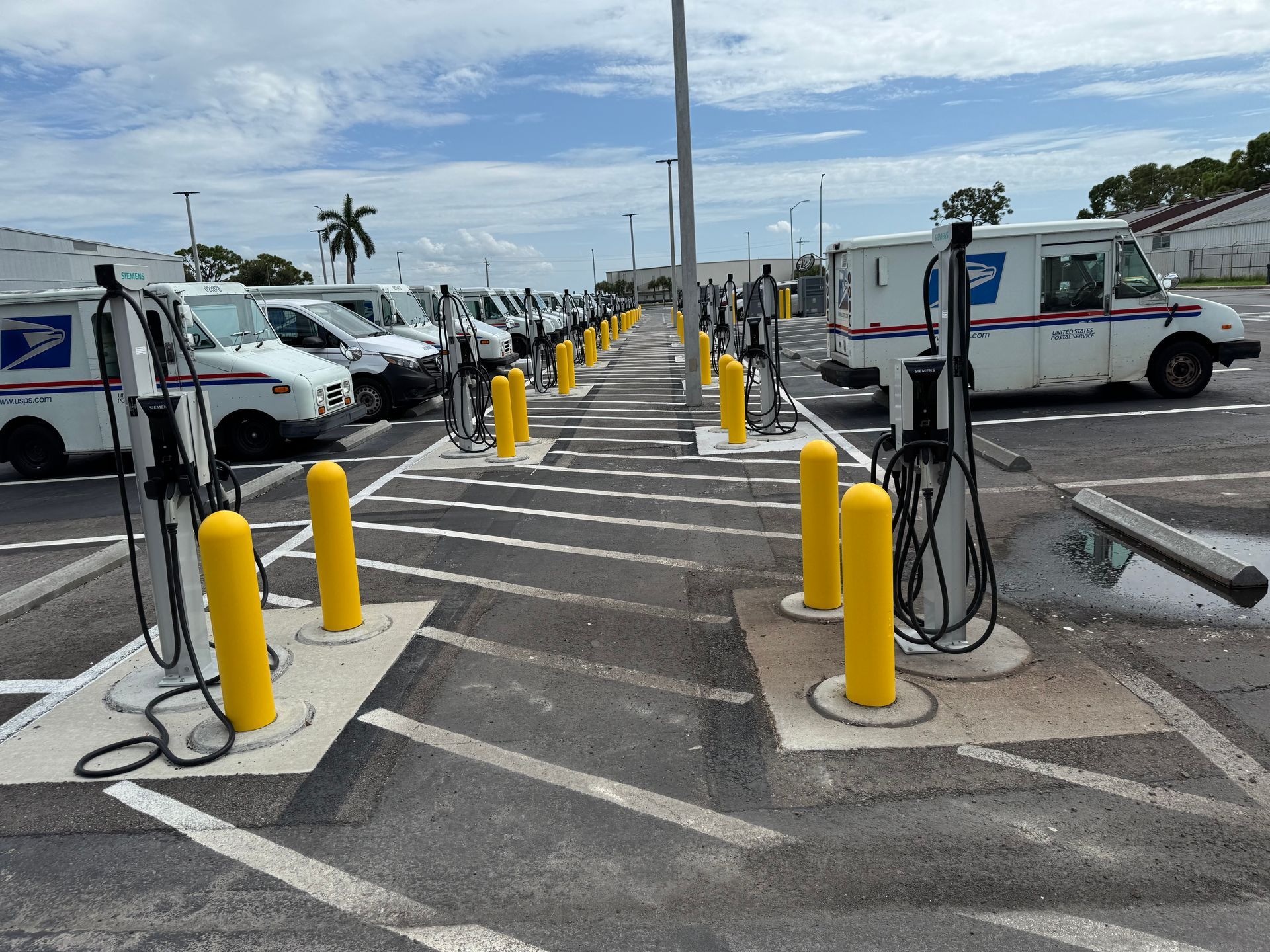 Postal service vans parked at charging stations. Yellow bollards and chargers. Outdoor setting.