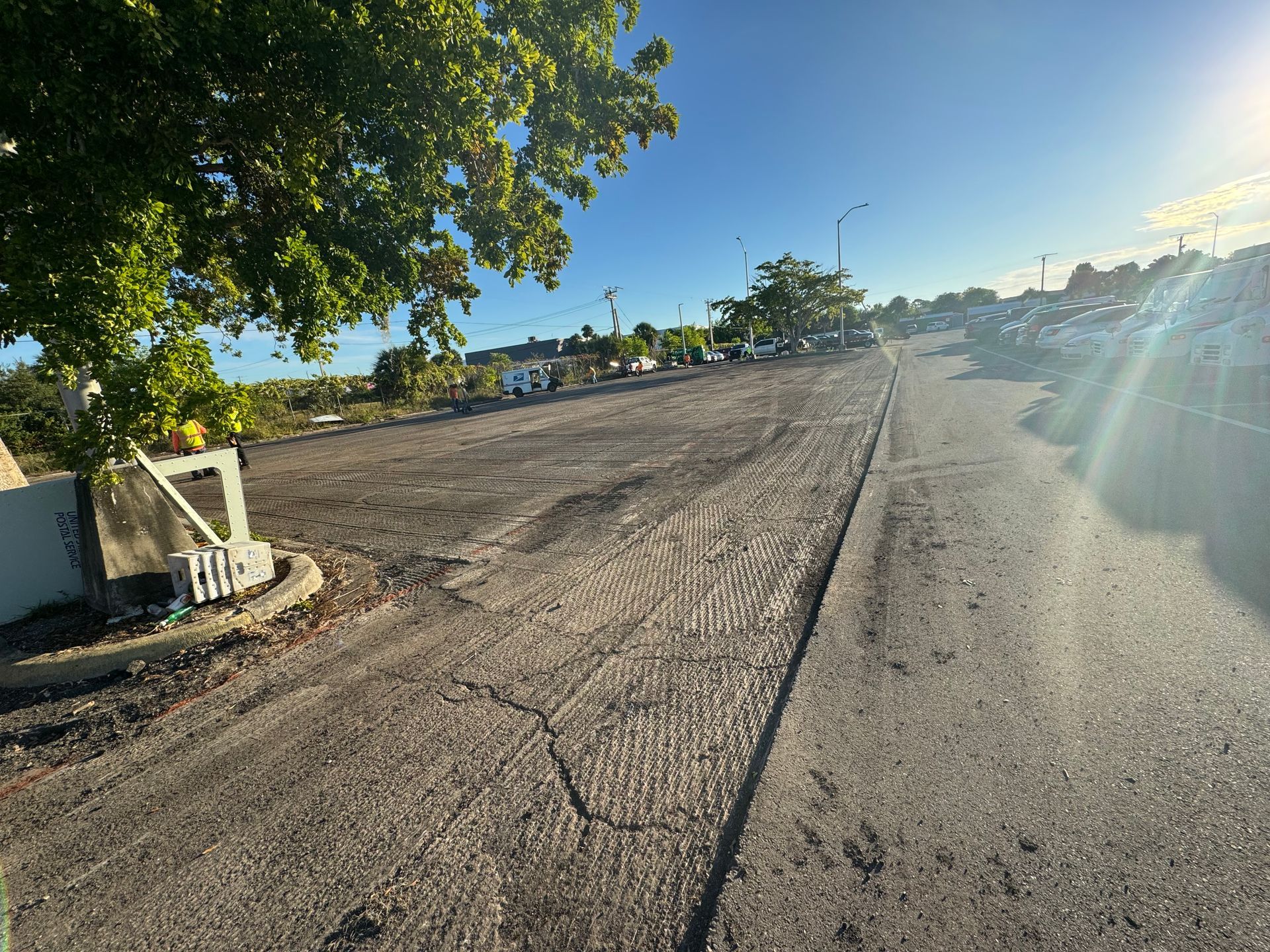 Asphalt road with cracked surface, leading to a parking area with vehicles. Bright sky, tree on the left.
