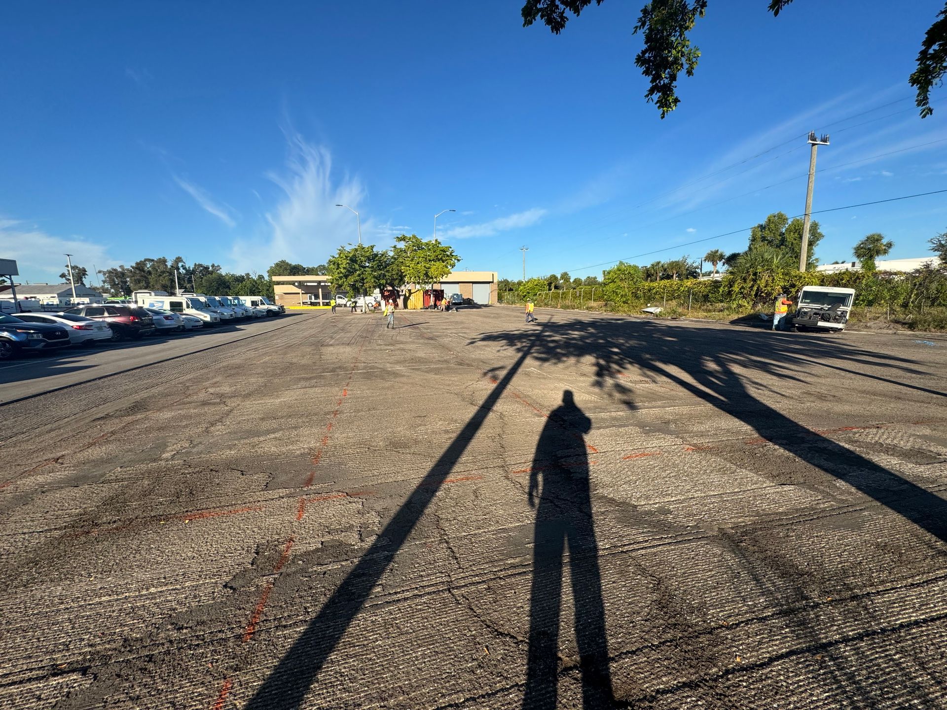 Open gravel lot under a clear, blue sky, with long shadow of person in foreground. Buildings and parked cars in background.