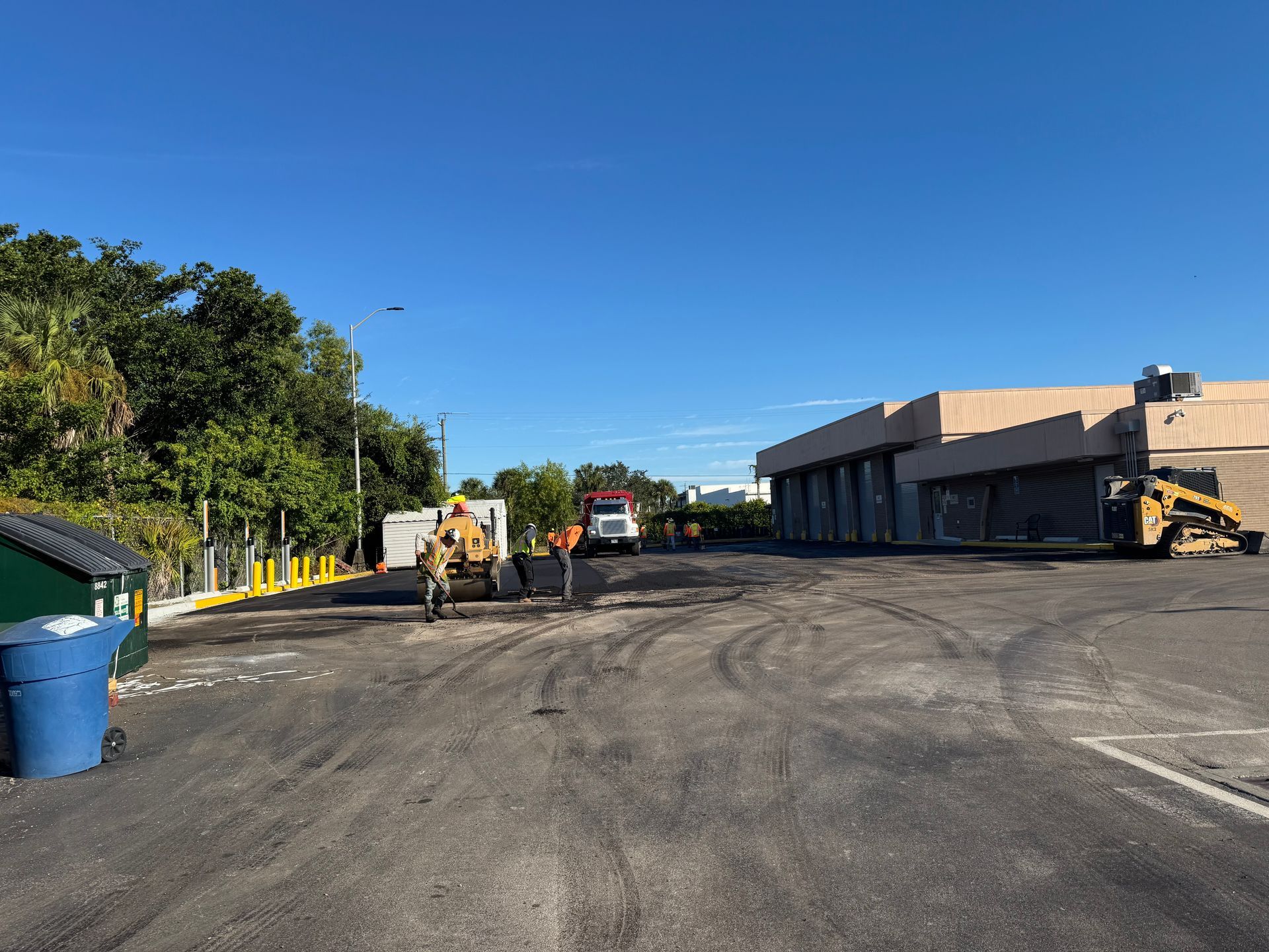 Construction site; workers, bulldozer, and truck. Building and trees in the background, blue sky.