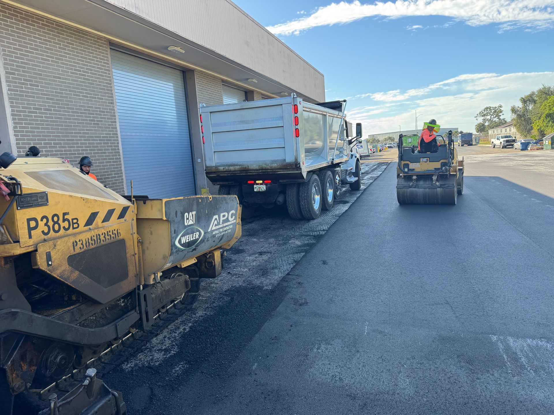 Asphalt paving: Dump truck, paving machine, and roller working on a road next to a building.