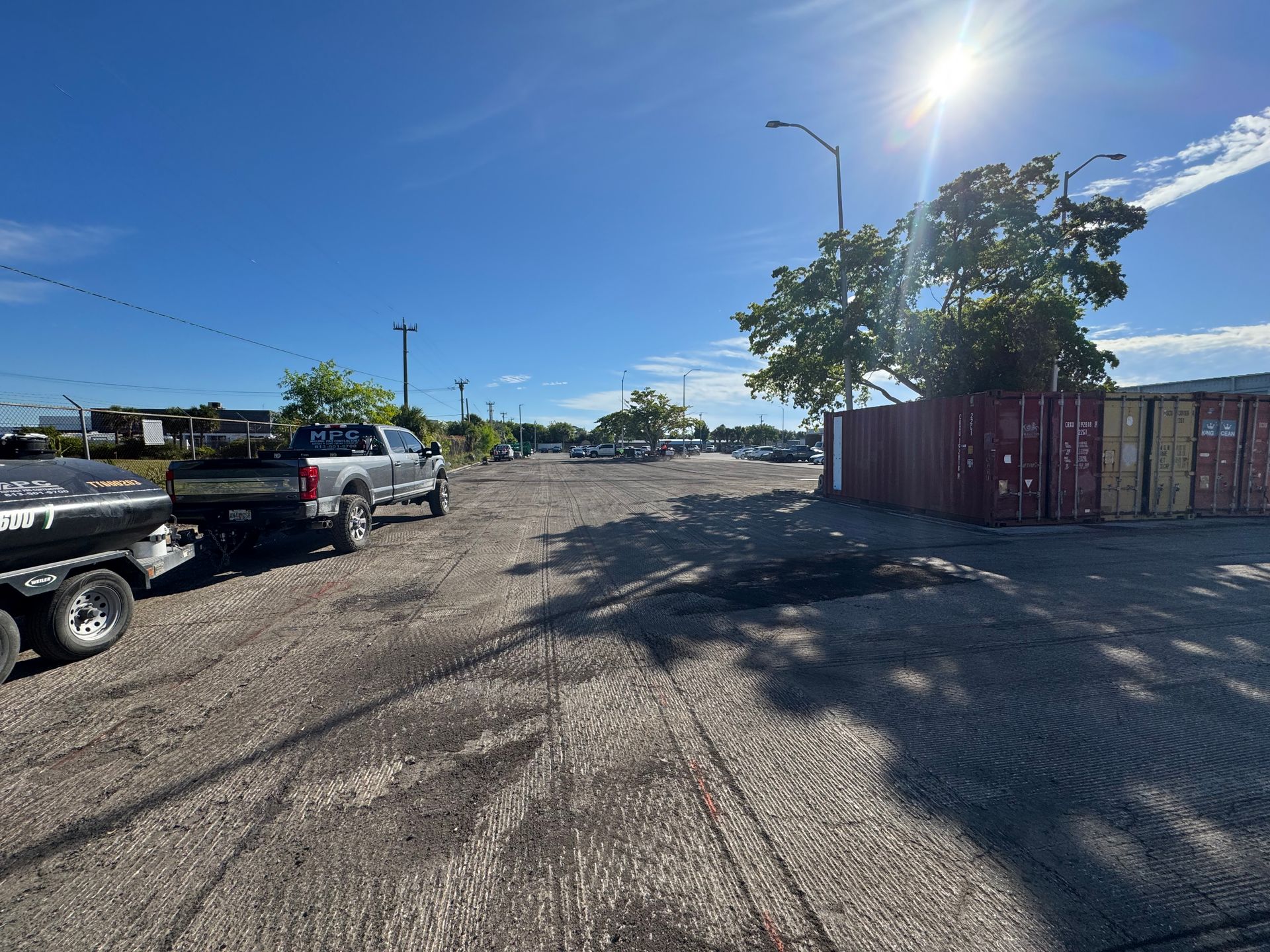 A gravel parking lot with a pickup truck and trailer under a bright sun. A shipping container fence is on the right.
