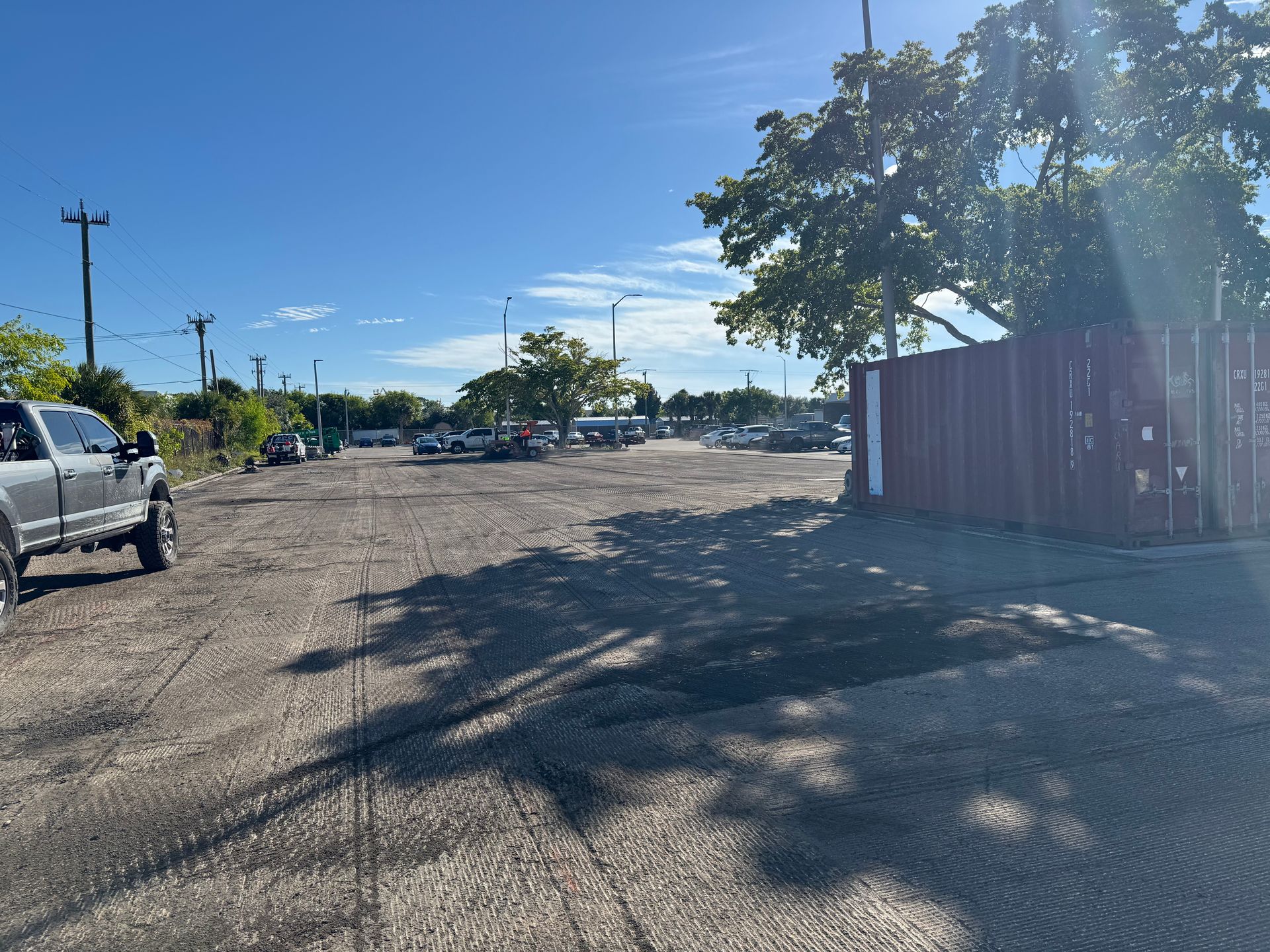 Gravel parking lot on a sunny day with cars, a truck, trees, and a red container.